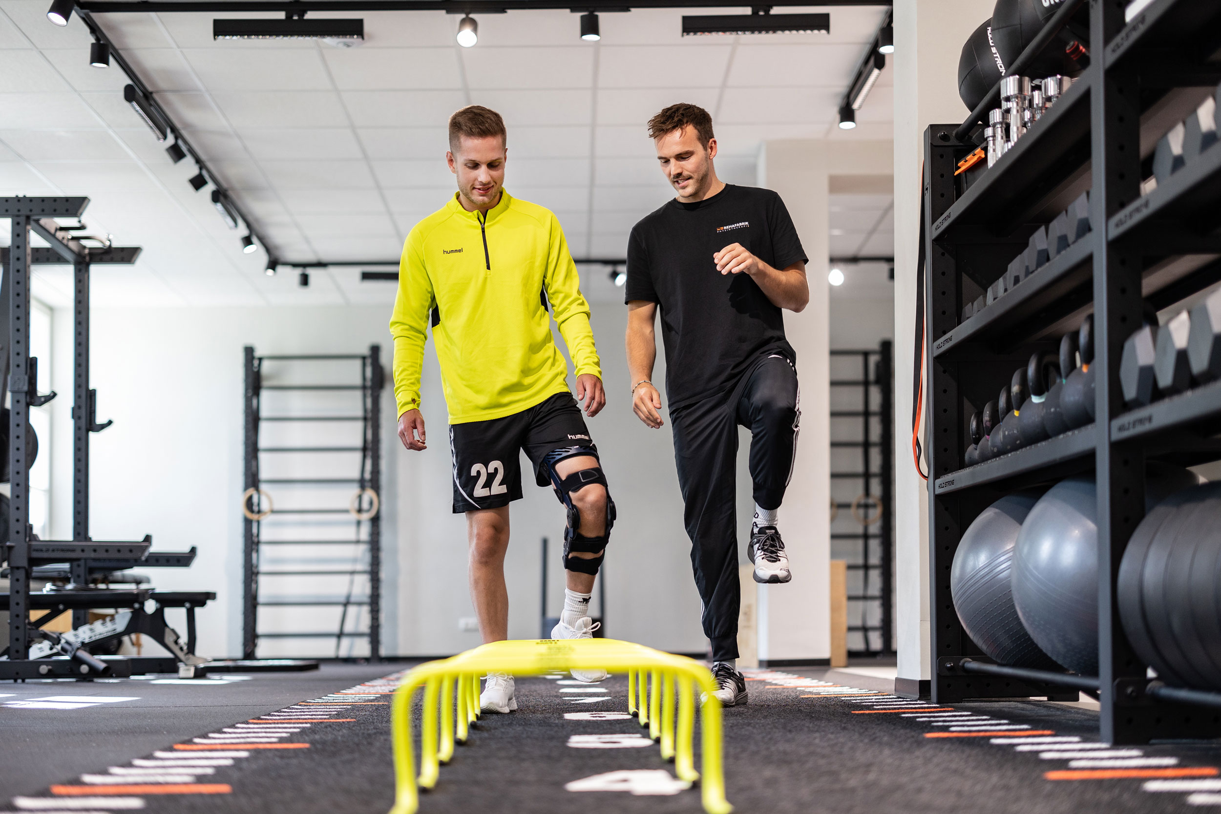 Eine Person mit gelbem Shirt macht Physiotherapie nach einer Knie-OP und absolviert ein Rehaprogramm über Hürden unter Anleitung eines Trainers mit schwarzem T-Shirt