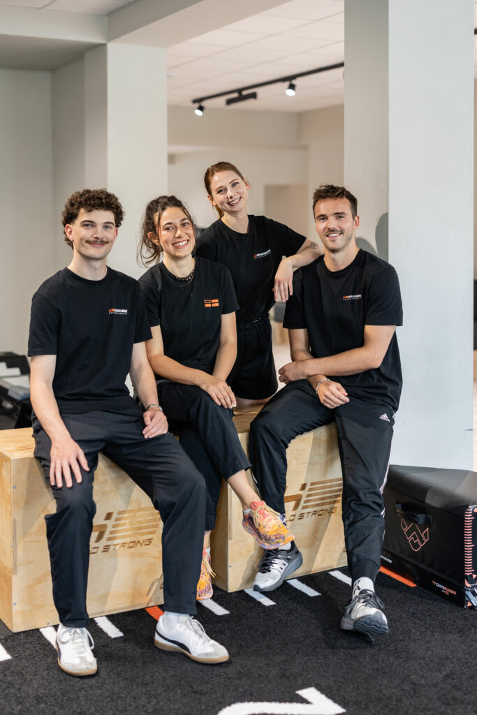 REHAFABRIK Physiotherapeut:innen zum Gruppenbild aufgestellt mit richtig guter Laune. Alle tragen eine schwarzes REHAFABRIK T-Shirt mit Logo der REHAFABRIK. Sie befinden sich auf der Trainngsfläche der Physiopraxis REHAFABRIK.