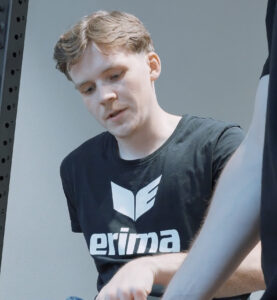 Junger Mann bei der Physiotherapie in der REHAFABRIK. Er trägt ein schwarzes T-Shirt mit einem weißen Aufdruck "erima". Dein Comeback mit REHAFABRIK .
