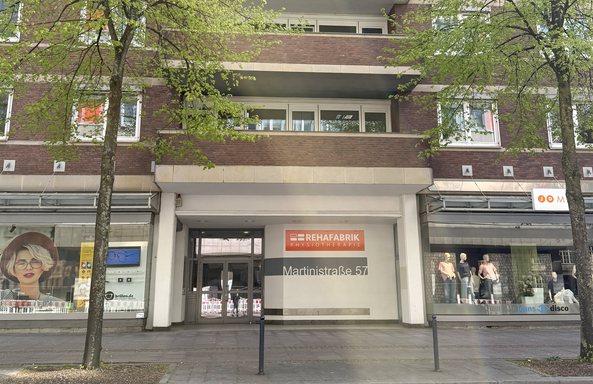 Der Eingang der REHAFABRIK Praxis in Bremen City am Brill. Vor dem Gebäude stehen Bäume. Am Gebäude befindet sich ein auffälliges orangenes Schild.